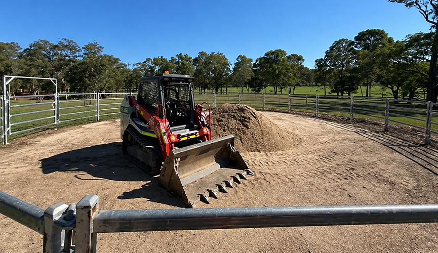A skid steer loader working on an outdoor arena during the horse ménage construction process. Horse arena builders are preparing the site for a small dressage arena, ensuring accurate horse arena measurements and stable arena footing. This equestrian riding arena project highlights the importance of site leveling and proper horse arena dimensions for training in smaller areas.