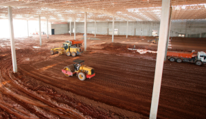 Professional equestrian arena builders performing horse arena construction inside a large indoor arena structure. The image shows heavy machinery, including a laser grading roller and a grader, leveling the base of a 20m x 60m standard dressage arena. This stage of equine arena construction ensures precise dressage arena dimensions and a stable foundation for the final arena footing.