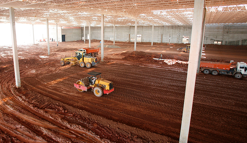 Professional equestrian arena builders performing horse arena construction inside a large indoor arena structure. The image shows heavy machinery, including a laser grading roller and a grader, leveling the base of a 20m x 60m standard dressage arena. This stage of equine arena construction ensures precise dressage arena dimensions and a stable foundation for the final arena footing.