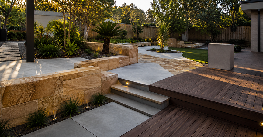 Modern backyard hardscaping featuring a dark timber deck, concrete steps with integrated lighting, sandstone block retaining walls, and a curved concrete and stone paved pathway leading through a landscaped garden.