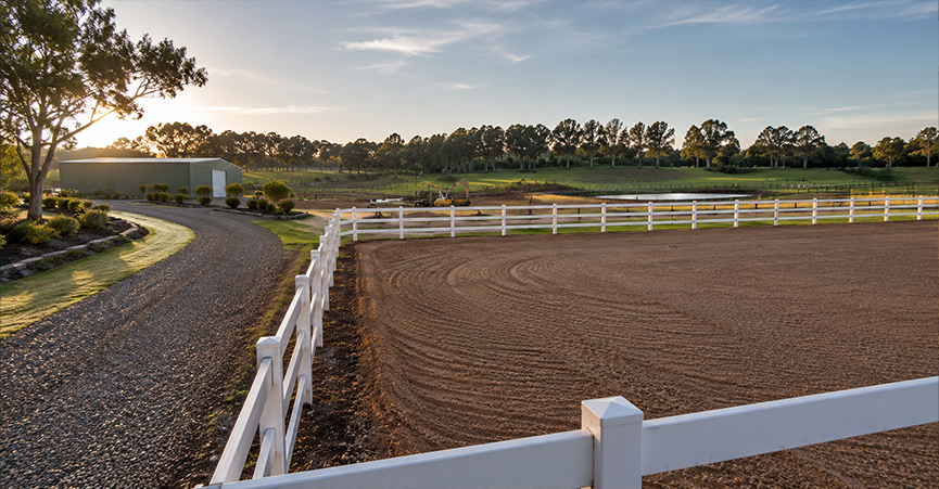 Specialized rural landscaping featuring a newly constructed sand horse arena with a white post-and-rail fence, a curved gravel driveway, an acreage farm dam, and a large green machinery shed in the background at sunset.